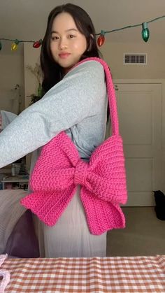 a woman holding a pink purse with a big bow on it's shoulder and standing in front of a table