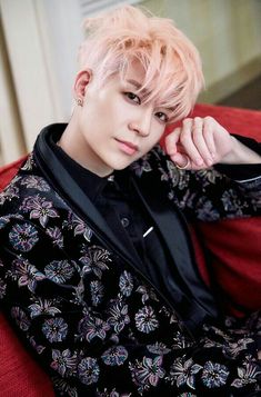 a young man with pink hair sitting on a red couch wearing a black shirt and floral pants