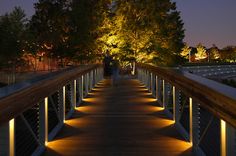 people walking across a bridge at night with lights on the walkway and trees in the background