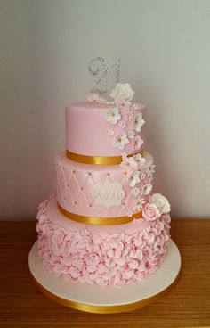 a three tiered pink cake with gold trim and flowers on the bottom, topped with a monogram