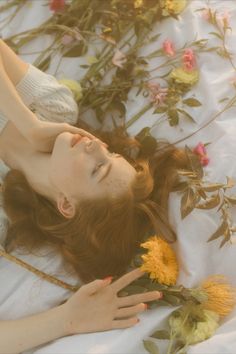 a woman laying on top of a white sheet next to yellow and pink flowers with her hands in the air