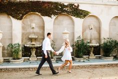 a man and woman holding hands walking in front of a wall with water fountains