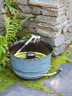 a garden hose is in a blue bucket next to a rock wall and green plants