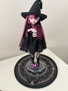 a doll with pink hair wearing a black dress and hat on top of a table