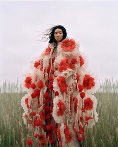 a woman standing in tall grass with red and white flowers on her cape over her shoulders