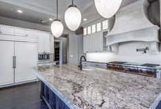 a large kitchen with marble counter tops and white cabinets, hanging lights over the stove