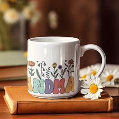 a white coffee mug sitting on top of a wooden table with daisies in front of it