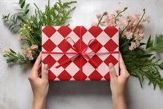 a person holding a red and white box with a bow on it, surrounded by flowers