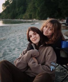 two young women sitting next to each other on the beach with trees in the background