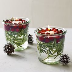 two glass cups filled with candles sitting on top of a table next to pine cones