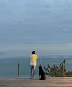 a man standing next to a black dog on top of a wooden deck near the ocean