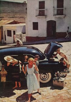 a woman standing in front of an old black car with luggage on the back and people looking at it