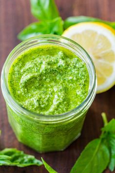 a jar filled with green pesto next to sliced lemons and basil on a wooden table