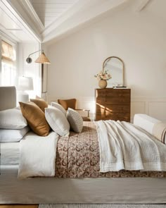 a bed with white sheets and pillows in a bedroom next to a dresser, mirror and lamp