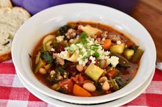 a white bowl filled with vegetable soup next to a piece of bread on a red and white checkered table cloth