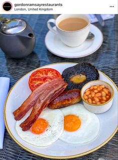 an english breakfast with bacon, eggs, tomatoes and beans on a plate next to a cup of coffee