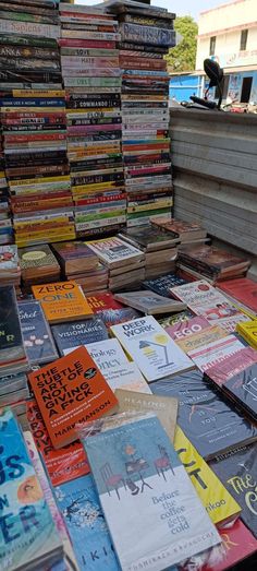 a pile of books sitting on top of a wooden table next to a building with people standing in the background