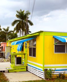 a small yellow house with a blue and white striped awning on the roof next to palm trees