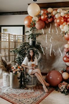 a woman sitting on a chair in front of a backdrop with balloons and greenery