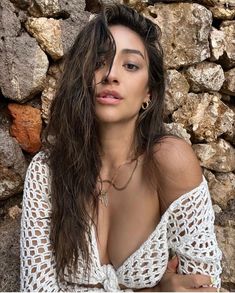 a woman with long hair posing in front of a stone wall wearing a white top