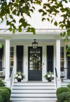 a white house with black shutters and two planters on the front steps, surrounded by greenery