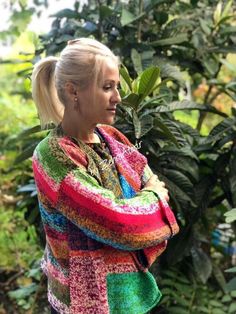 a woman with blonde hair wearing a multicolored cardigan standing in front of trees