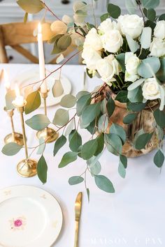 the table is set with white roses and greenery, gold place settings and candles