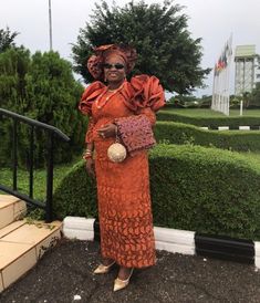 a woman in an orange dress standing next to some bushes and stairs with a purse on her shoulder