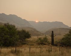 the sun is setting in the mountains behind a fenced off field with trees and bushes