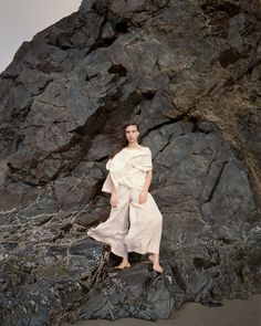 a woman standing on top of a rocky beach next to a large rock covered hillside