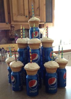 a stack of pepsi cans with cupcakes and candles in them on a kitchen counter