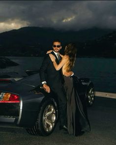 a man and woman standing next to a black sports car in front of the ocean