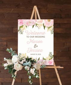 a welcome sign with flowers and greenery sits on an easel in front of a wooden wall