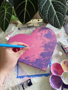 a person is painting a heart with pink and purple paint on paper next to some plants