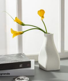 a white vase with yellow flowers sitting on top of a table next to a book