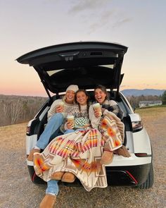 two women sitting in the back of a car with their blankets on and holding coffee cups