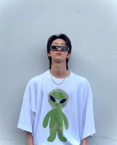 a young man wearing sunglasses and an alien t - shirt stands against a white wall