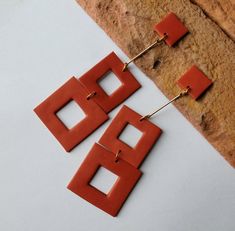 two square shaped orange earrings on top of a piece of paper next to a rock