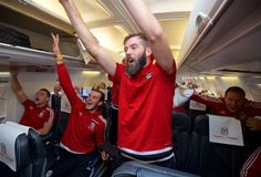 two men in red shirts on an airplane with their arms up and one man holding his hands up