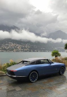 a blue sports car parked next to a body of water with mountains in the background