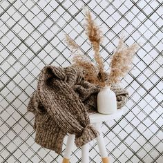 a knitted hat and scarf sitting on a stool next to a vase with dry grass in it