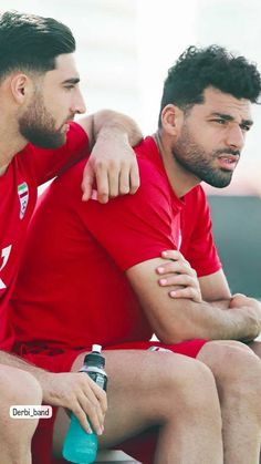 two men sitting next to each other wearing red shirts and shorts, one with his arm around the other's shoulder