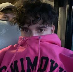 a young man with curly hair wearing a pink hoodie and looking at the camera