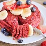 a white plate topped with red velvet pancakes covered in bananas and blueberries next to strawberries