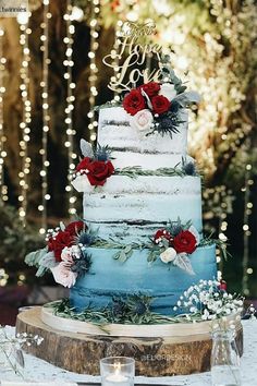 a blue and white wedding cake with red flowers on the top is surrounded by string lights