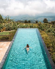 a woman swimming in a large pool surrounded by greenery and mountains on a cloudy day