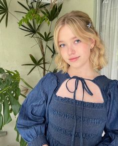 a woman in a blue dress standing next to a potted plant and looking at the camera