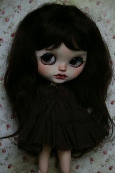 a doll with long black hair and big eyes wearing a dark dress on a floral wallpapered background