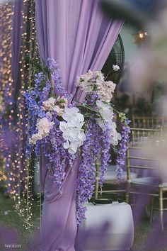 purple and white flowers are hanging from the back of a wedding ceremony arch with draping