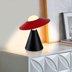 a red and black table lamp sitting on top of a white dresser next to a framed photograph